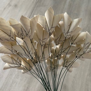 May include: A bouquet of paper flowers made from book pages. The flowers are white and have a delicate, vintage look. The stems are green wire.