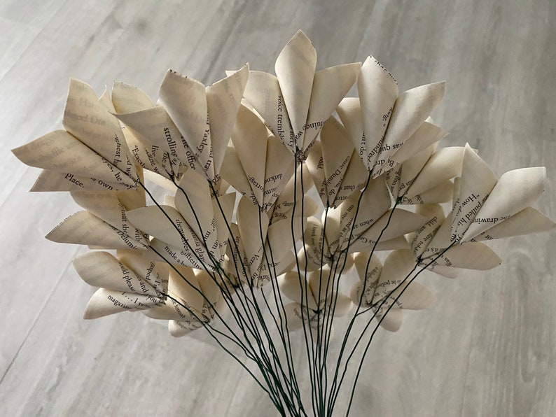 May include: A bouquet of paper flowers made from book pages. The flowers are white and have a delicate, vintage look. The stems are green wire.