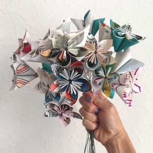 May include: A bouquet of folded paper flowers crafted from recycled book pages and colorful printed paper. The individual blossoms feature various geometric shapes, patterns, and text, with shades of teal, red, black, and white visible throughout.