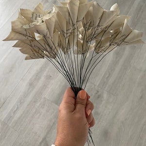 May include: A bouquet of paper flowers made from the pages of a book. The flowers are white and have a rustic, vintage look. The stems are made of wire and are held together by a hand.