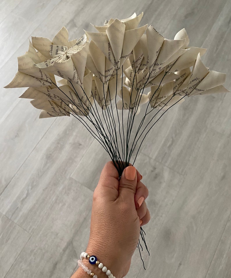 May include: A bouquet of paper flowers made from the pages of a book. The flowers are white and have a rustic, vintage look. The stems are made of wire and are held together by a hand.