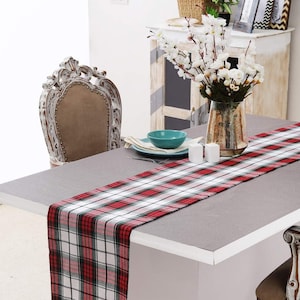 May include: A dining table set with a red, black, and white plaid table runner. The table is set with a teal bowl, plates, and candles. A decorative chair is on the side. A vase of white flowers sits in the background.