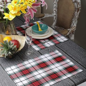 May include: Two rectangular placemats with a red, green, black, and white plaid pattern are on a gray wooden table. A bowl, plates, and a glass are on top of the placemats. A vase of flowers and a bowl of fruit are also on the table.