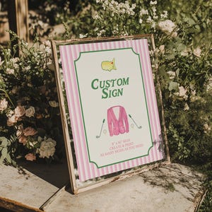 Puede incluir: Un letrero personalizado enmarcado con un fondo a rayas rosas y blancas. El letrero presenta el texto "Custom Sign" en verde, junto con un gr&aacute;fico de chaqueta rosa y palos de golf. El letrero mide 20,3 cm x 25,4 cm y est&aacute; rodeado de vegetaci&oacute;n y flores.