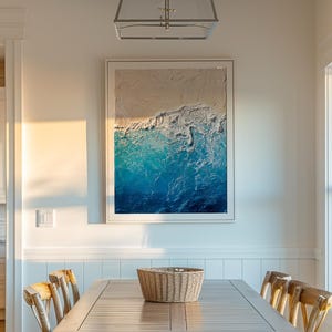 May include: A rectangular framed wall art piece featuring a textured abstract ocean scene with white sand and gradient blue water, hanging above a wooden dining table with four chairs and a woven basket in a bright, sunlit room.