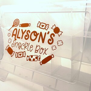 May include: A clear plastic snack box with multiple compartments. The front of the box is decorated with brown candy illustrations and the text "ALYSON'S Snack Box". The box is designed to hold various snacks.