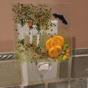 May include: A decorative night light featuring a glass panel with a fall theme. The panel depicts a tree with green and orange foliage, two orange pumpkins, and a black bat. The night light is white and plugs into a wall outlet.