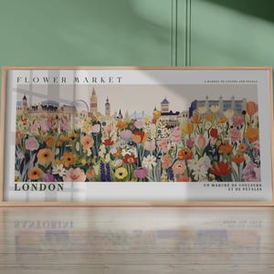 以下が含まれることがあります： ロンドンのスカイラインを背景にした、カラフルな花のイラストが描かれた額入りのアートプリント。「FLOWER MARKET」と「LONDON」の文字とフランス語のテキストが含まれています。ピンク、オレンジ、黄色、紫の色合いの花々が描かれたアートワークです。