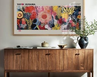 May include: A framed art print featuring a colorful floral design by Yayoi Kusama hangs above a wooden cabinet. The cabinet has four doors with dark wooden handles. A vase with greenery, a bowl, and books are on top of the cabinet.