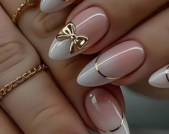 May include: Oval-shaped nails with a white and pink ombre design, accented with gold trim and a gold bow. The nails are long and well-manicured, showcasing a delicate and elegant aesthetic. The overall look is sophisticated and stylish.
