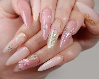 Peut inclure: Une main avec de longs ongles en acrylique pointus, roses et transparents. Certains ongles présentent des motifs floraux délicats roses et blancs, rehaussés de bordures dorées. D'autres sont roses unis ou transparents avec des détails de gouttes d'eau.
