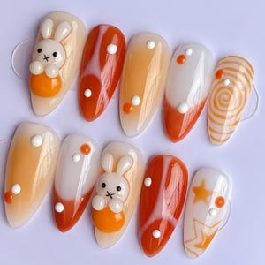 May include: Set of ten false nails with an orange and white color scheme. Some nails feature a swirl design, others have white dots, and some have a 3D bunny charm.