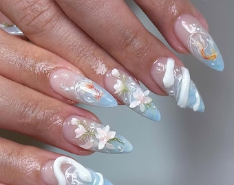 May include: Close-up of a hand with light blue, long, pointed nails decorated with white 3D flowers, a white snake, and a small orange fish. The nails are decorated with a clear gel and have a light blue base.