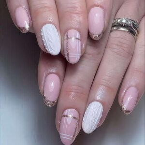 May include: A set of manicured nails featuring a variety of designs. Some nails are painted a pale pink with a gold glitter French tip. Other nails have a white cable knit design or a pink base with white lines and gold glitter accents.