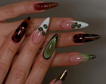 Puede incluir: Primer plano de uñas largas y en forma de almendra con una variedad de diseños. Algunas uñas están pintadas con un patrón de caparazón de tortuga marrón y negro, mientras que otras presentan una base blanca con acentos verdes.