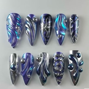 Peut inclure: Dix pointes d'ongles violettes et argentées avec des motifs abstraits différents. Chaque pointe d'ongle a un accent argenté et un petit bijou transparent.