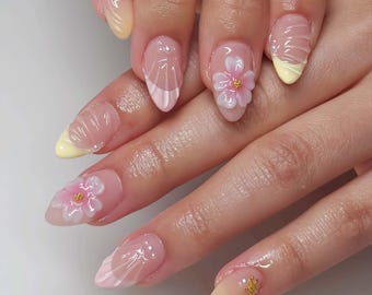 May include: Close-up of a hand with almond-shaped nails. The nails feature a clear base with various designs, including a white and pink French tip, 3D flower accents, and gold star embellishments. Some nails have a yellow tip.
