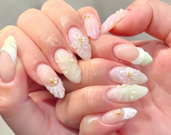 May include: A hand displaying long, oval-shaped nails with diverse designs. Some nails have a French tip with a pastel green accent, while others feature 3D floral and seashell embellishments. The colour scheme includes soft pinks, whites, and gold accents.