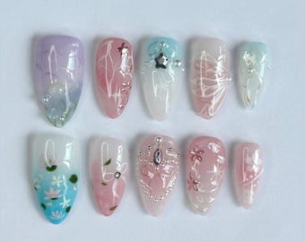 May include: A set of ten artificial nails in various shapes and sizes, each with a unique design. The nails feature pastel colors like pink, blue, and purple, adorned with floral patterns, gems, and glitter accents. The designs vary from abstract swirls to detailed floral art.