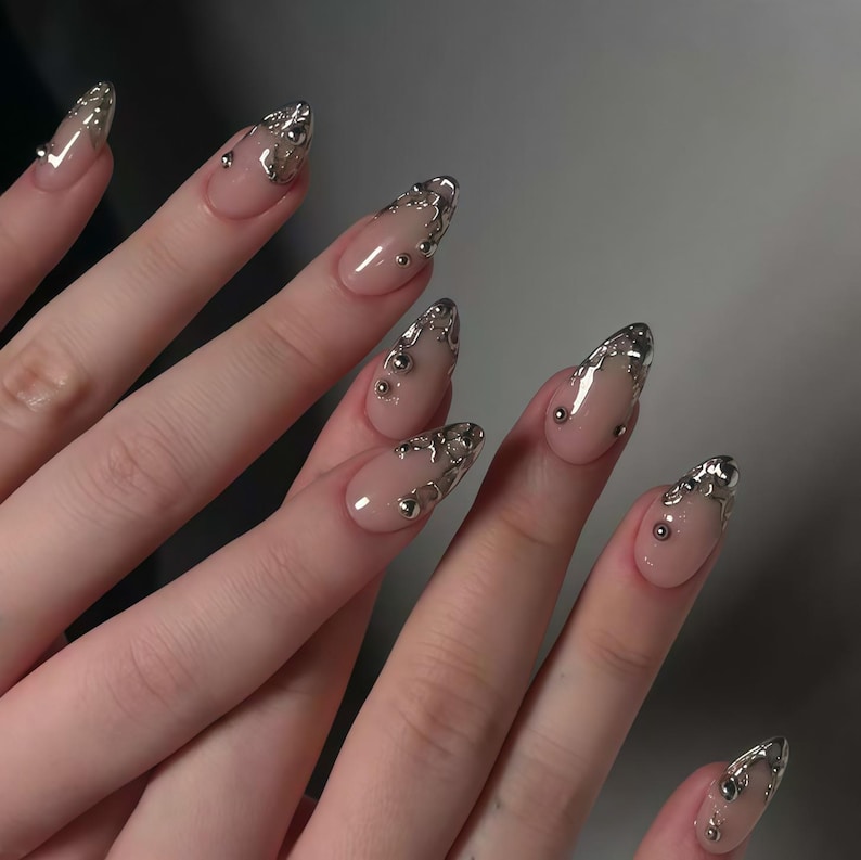 May include: Close-up of a hand with long, almond-shaped nails. The nails are painted a nude color with silver metallic accents along the tips and small silver beads. The design is intricate and eye-catching, perfect for a special occasion.
