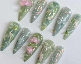 May include: Ten long, light green, almond-shaped false nails with intricate designs. Some nails feature a pink flower, while others have a white flower or a white fish. The nails have a glossy finish.