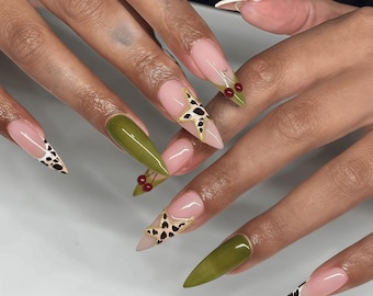 Puede incluir: Uñas acrílicas largas y puntiagudas. Las uñas presentan una variedad de diseños, incluyendo verde oliva liso, rosa con un patrón blanco y negro, y rosa con una estrella dorada y estampado de leopardo. Algunas uñas tienen pequeños detalles de cerezas.