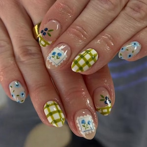May include: Close-up of manicured nails with various designs. Some nails feature a clear base with blue floral patterns, while others have green and white checkered patterns. Two nails display a blue floral stamp design.