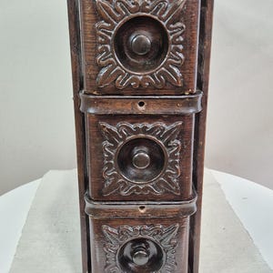 May include: A dark brown, three-drawer wooden cabinet with ornate floral carvings. Each drawer has a central knob and keyhole. The cabinet is vertically oriented and appears to be antique or vintage.