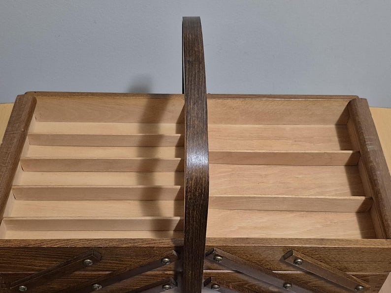 May include: A wooden sewing basket with a dark brown arched handle. The interior features multiple compartments and a hinged base. The basket is made of light and dark brown wood and has metal hardware.