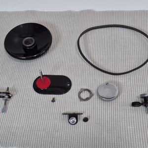 Peut inclure: Ensemble de pièces de platine vinyle vintage, comprenant une poulie noire, une courroie en caoutchouc, une plaque métallique avec un disque rouge et diverses vis et rondelles. Les composants sont disposés sur une surface texturée de couleur claire.