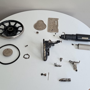 May include: Disassembled vintage Singer sewing machine parts, including a black hand wheel, a Singer-branded motor housing, and various metal components. The parts are arranged on a white surface, showcasing the intricate mechanics.