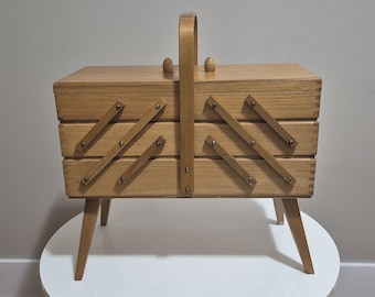 May include: A light-colored wooden sewing box with a handle and angled legs. The box has a tiered design with angled wooden supports and small metal rivets. The lid has a small wooden knob. The box is likely used for storing sewing supplies.