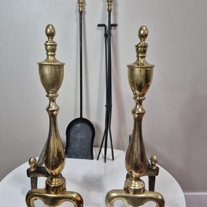 Peut inclure: Deux chenets en laiton avec un design d'urne décorative, reposant sur des pieds incurvés. Un ensemble d'outils de cheminée, dont une pelle, un tisonnier et des pinces, est visible en arrière-plan. Les outils ont des poignées noires et des accents dorés.