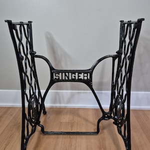 May include: A black cast iron sewing machine base featuring an intricate, openwork scroll design on the side supports. The central horizontal crossbar displays the word SINGER in raised, capital letters, connecting the two side frames.