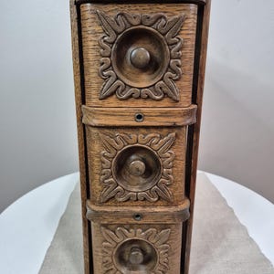 May include: A vertical wooden storage unit featuring three stacked drawers. Each drawer front is decorated with a carved square frame surrounding a circular center and a small round knob. The wood has a medium brown tone with visible grain.