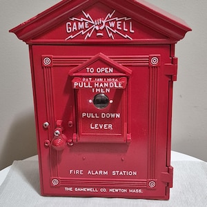 May include: A vintage red Gamewell fire alarm station. The box has the text "GAME WELL" and "FIRE ALARM STATION". It features a pull handle and lever, with instructions "TO OPEN" and "PULL DOWN LEVER". A set of keys is in front of the box.