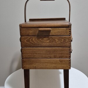 May include: A vintage wooden sewing box with a handle and four drawers. The box is made of dark brown wood and has a rectangular shape. It stands on four legs and is placed on a white surface.