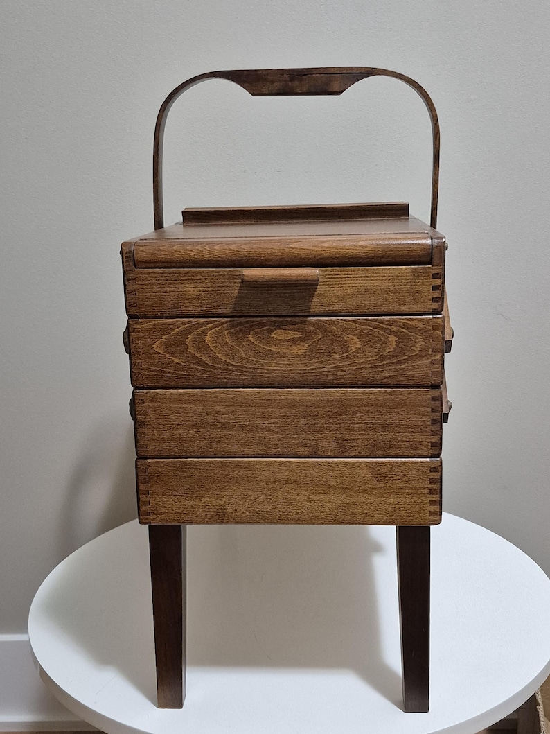 May include: A vintage wooden sewing box with a handle and four drawers. The box is made of dark brown wood and has a rectangular shape. It stands on four legs and is placed on a white surface.