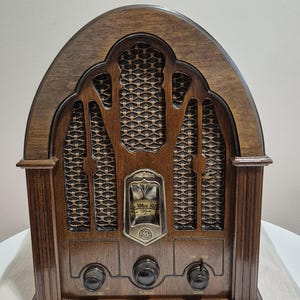 May include: A classic wooden radio in a dark brown finish. The radio has an arched top, a speaker grille, and control knobs for volume, tuning, and tone. The radio features an FM/AM dial and a GE logo.