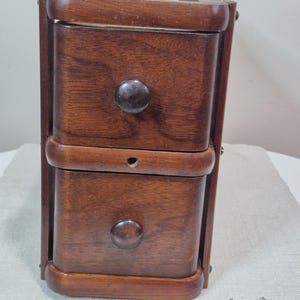 Peut inclure: Petit meuble en bois à deux tiroirs avec boutons marron foncé. Le meuble est en bois teinté et de forme rectangulaire. Quatre vis sont visibles en bas à droite. Le meuble est probablement utilisé pour le rangement.