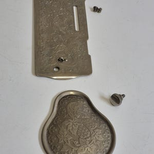 May include: Vintage sewing machine parts in silver. The image shows a rectangular plate with a slot and floral pattern, a pear-shaped plate with a grape and leaf design, and screws. These are likely antique components.