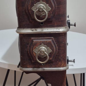 May include: A vintage, dark wood cabinet with two drawers. Each drawer has a decorative silver pull with a ring handle. The cabinet has a silver trim and is mounted on a small wooden shelf.