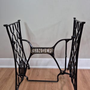 May include: A black cast iron Singer sewing machine treadle base featuring an intricate, openwork scroll design on the side panels and a central horizontal brace prominently displaying the word SINGER in raised, light-colored lettering.
