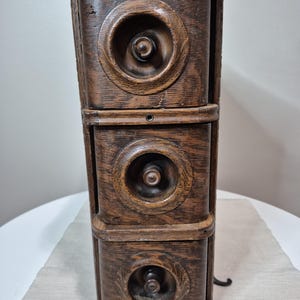 Peut inclure: Un meuble de machine à coudre ancien en bois avec trois tiroirs. Chaque tiroir présente un élément décoratif circulaire avec un bouton central. Le meuble est en bois teinté foncé, mettant en valeur le grain naturel. Le design général suggère une esthétique classique et antique.