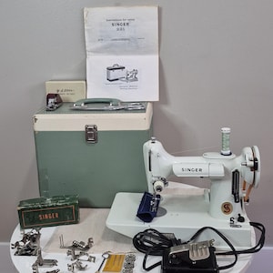 May include: A vintage white Singer 221 Featherweight sewing machine with a green carrying case, instruction manual, black foot pedal, and various metal presser feet, bobbins, and needles arranged on a white surface with a green Singer box.