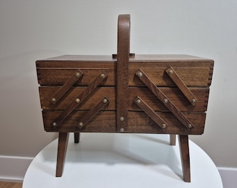 May include: A vintage wooden sewing box with a handle and angled supports. The box is made of dark brown wood and features brass-colored hardware. The box has a tiered design with angled supports and a handle for carrying.