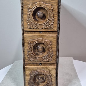 May include: A vertical wooden storage unit featuring three stacked drawers. Each drawer front displays a circular carved relief pattern with a central knob. The wood has a medium brown finish with visible grain and a beaded trim along the edges.