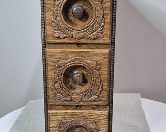 May include: A vertical wooden storage unit featuring three stacked drawers. Each drawer front displays a circular carved relief pattern with a central knob. The wood has a medium brown finish with visible grain and a beaded trim along the edges.