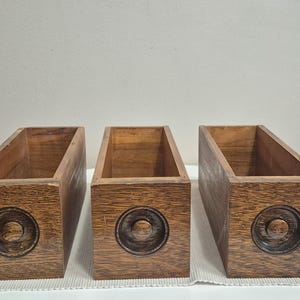 May include: Three rectangular wooden boxes in a natural wood finish. Each box features a carved circular design on the front. The boxes are open at the top and appear to be empty.