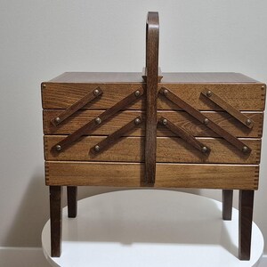 May include: A vintage wooden sewing box with a handle, featuring a multi-tiered design that expands outwards. The box is made of light brown wood, with brass-colored hardware and sits on four short legs. The handle is arched and made of the same wood.
